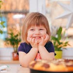 A happy young girl smiling indoors surrounded by plants and a pastry, with natural light streaming in.