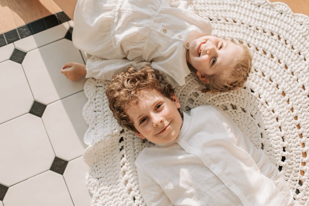 Two smiling siblings in white outfits lie on a woven rug in a bright room.