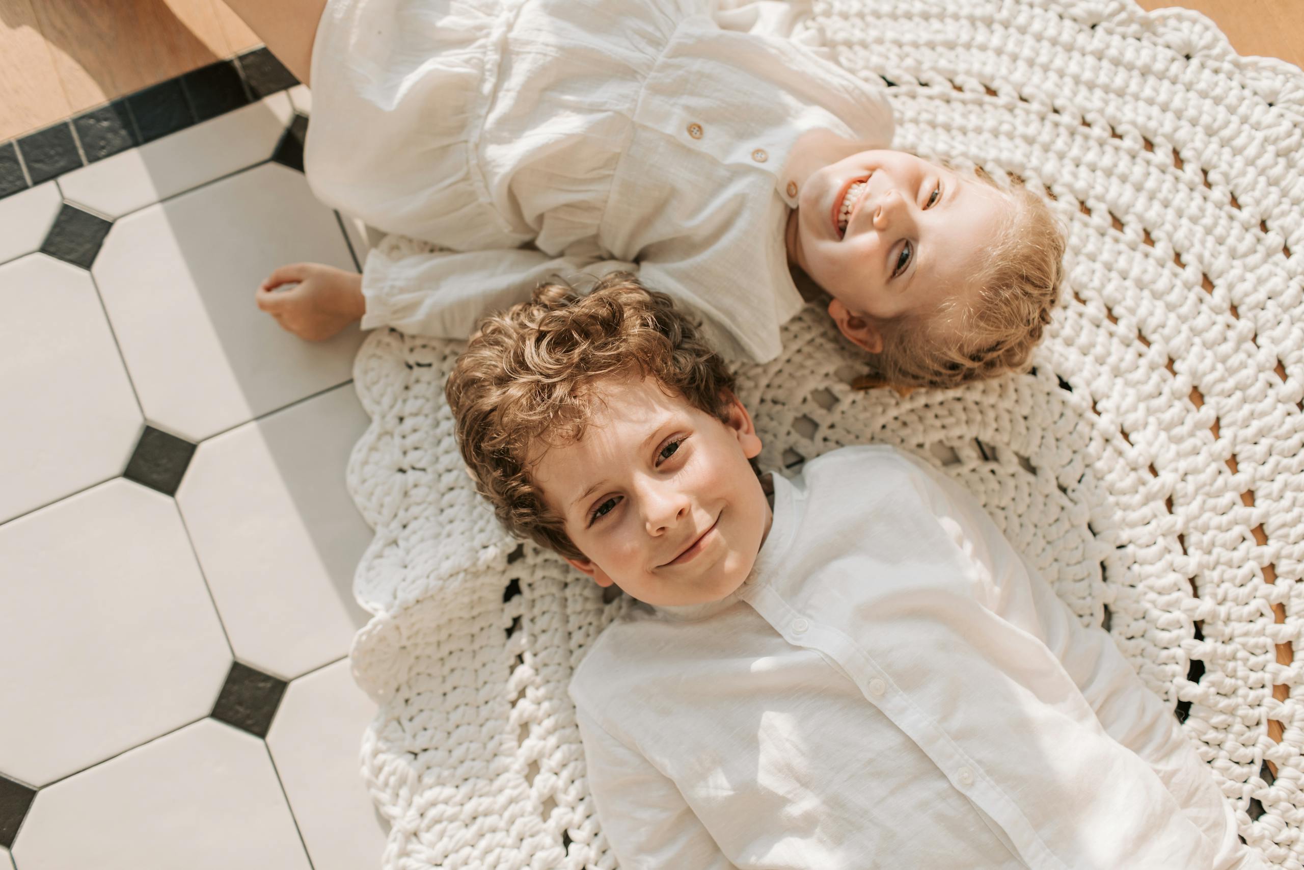 Two smiling siblings in white outfits lie on a woven rug in a bright room.